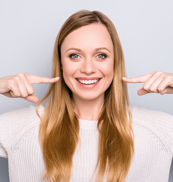 Smiling woman with long brown hair pointing to her teeth against white background.