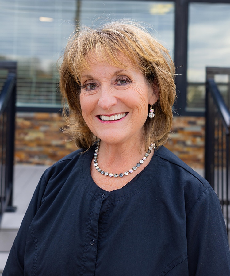 Professional woman smiling at camera in modern dental office setting with windows.