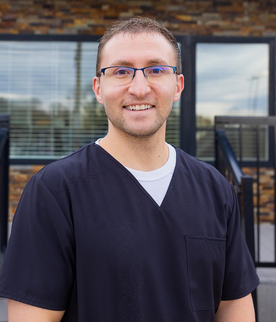 Male dental professional wearing glasses and dark scrubs smiling in front of modern office windows.