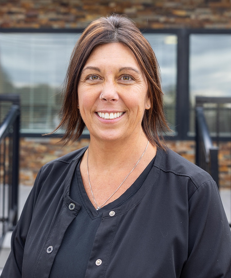 Professional woman smiling at camera wearing black scrubs in front of dental office building.