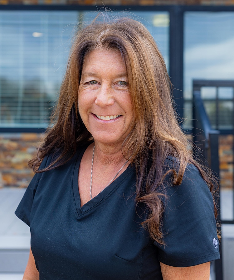 Woman smiling at camera wearing dark scrubs in front of modern dental office building.