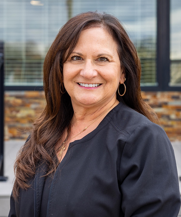Professional woman smiling at camera in dental office setting wearing black scrubs.