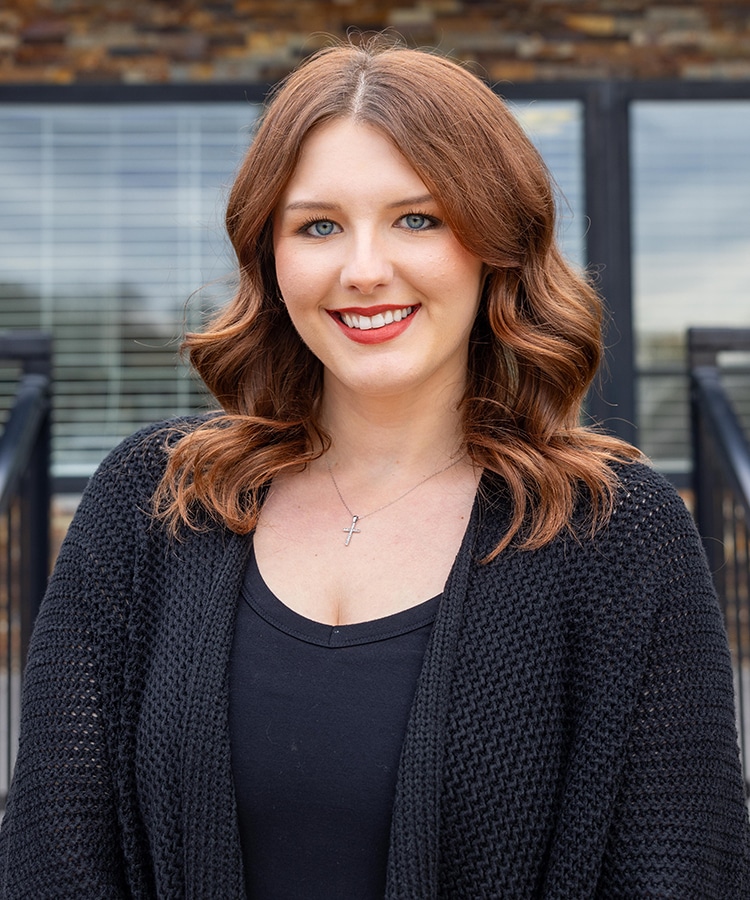 Professional woman with long brown wavy hair smiling at camera wearing dark cardigan against modern building backdrop.
