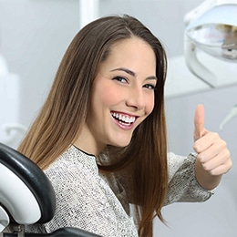 Happy woman giving thumbs up in dental office setting with dental equipment visible.