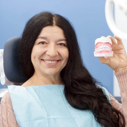 Woman in dental chair holding dental product, smiling at camera in modern dental office setting.