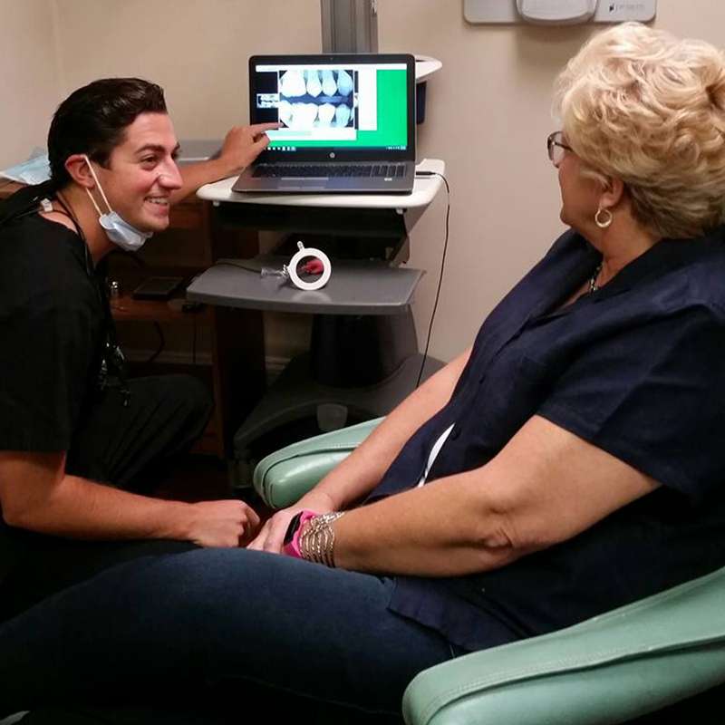 Dentist examining patient's arm while displaying dental X-rays on monitor in clinical setting.