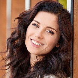 Smiling woman with long dark wavy hair in front of wooden door background for dental practice.