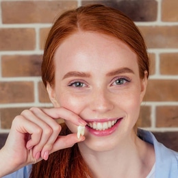 Smiling woman with red hair holding dental floss against brick wall background.