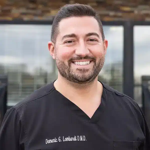 Male dentist in black scrubs smiling outdoors with modern building behind him.
