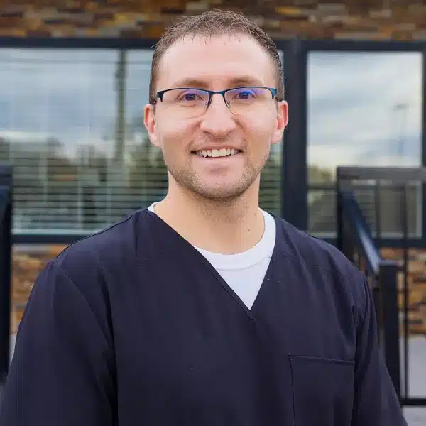 Young man with glasses smiling at camera in modern dental office setting.